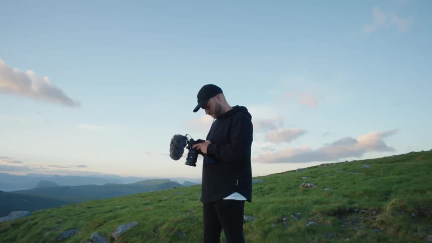 A man is standing outside on a grass covered hill looking at his camera and there are mountains in the distance