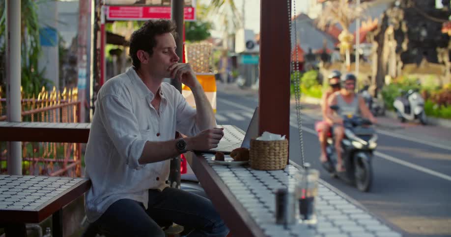 A man is sitting at an outdoor cafe typing on a laptop computer as traffic moves by on the street
