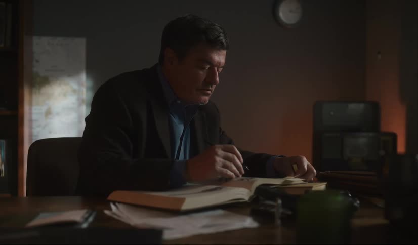 A man sits with a book and uses pen to write something on the page hes looking at
