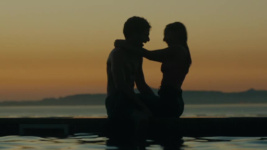 Sitting couple silhouetted against the red sky hug and laugh while sitting near pool with ocean in background