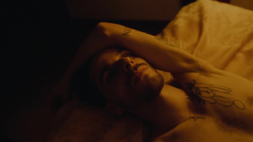 A young man with tattoos lays on his back in bed while staring off into the distance