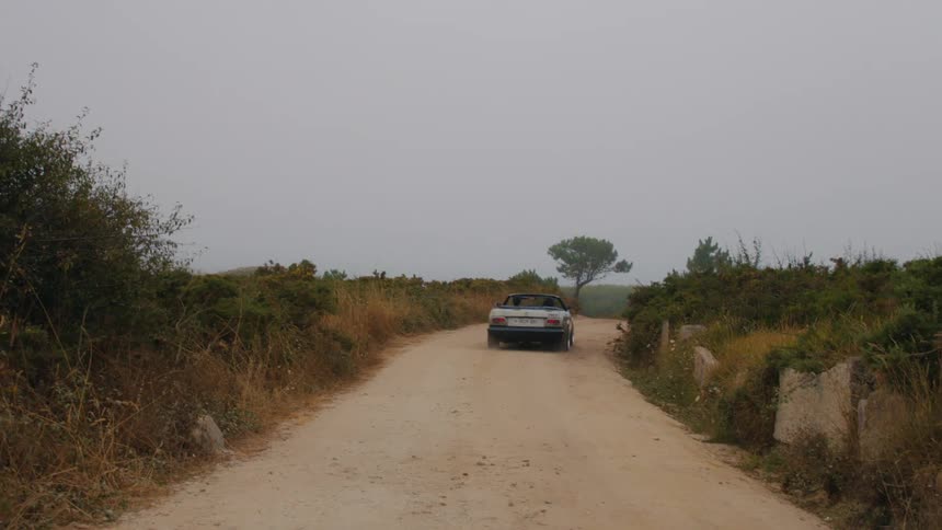 Two people drive along dirt road in convertible on an overcast day