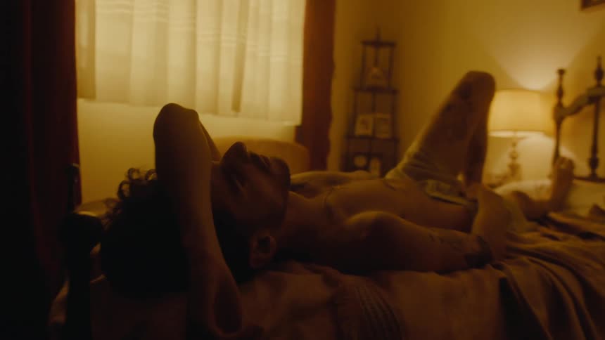 A young man lays shirtless on a bed with his arm across his forehead