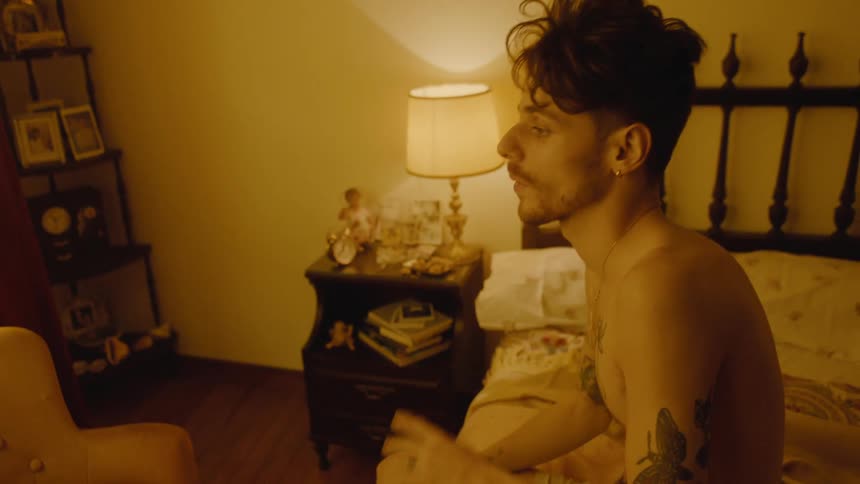 A young man with tattoos slowly rolls over in bed to get up and look out the window