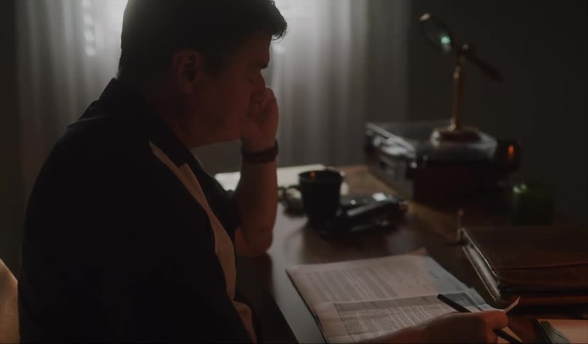Man with dark shirt and wristwatch sitting at desk holds paper while talking on smartphone in room
