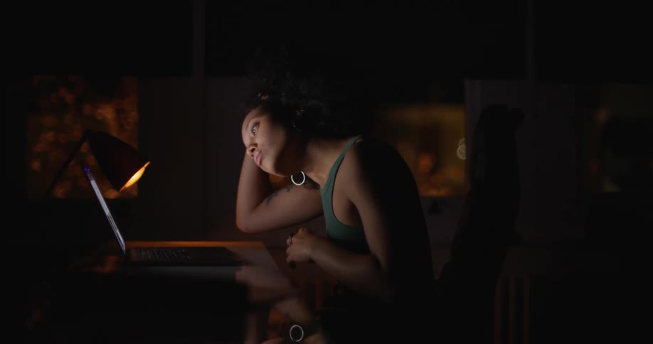 Woman with curly hair earrings and green top sits at desk working on laptop in dark room