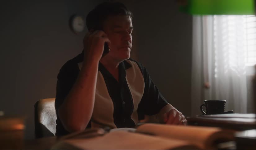 A man sitting at a desk writing in a book pauses to answer his phone