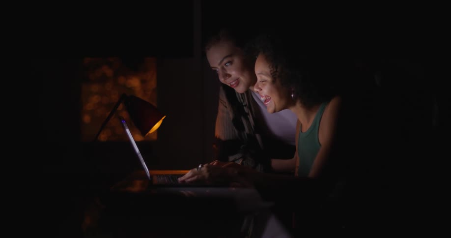 Two women laugh as they work on a laptop at night.