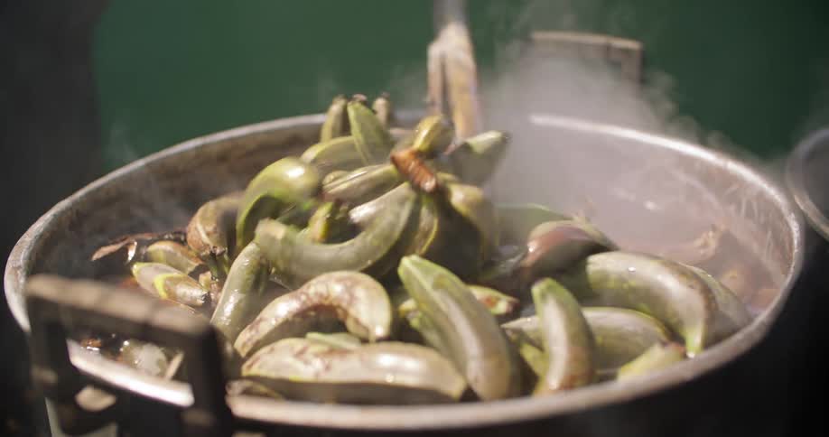 A person uses a long stick to stir a pot of boiling bananas