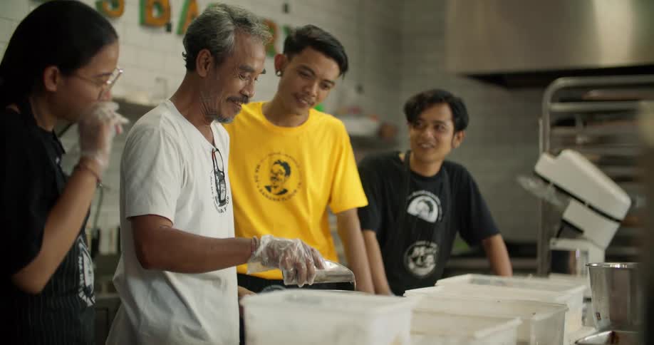A middle aged man scoops ingredients from a plastic container and pours them into a pot while speaking with the younger people watching from beside him