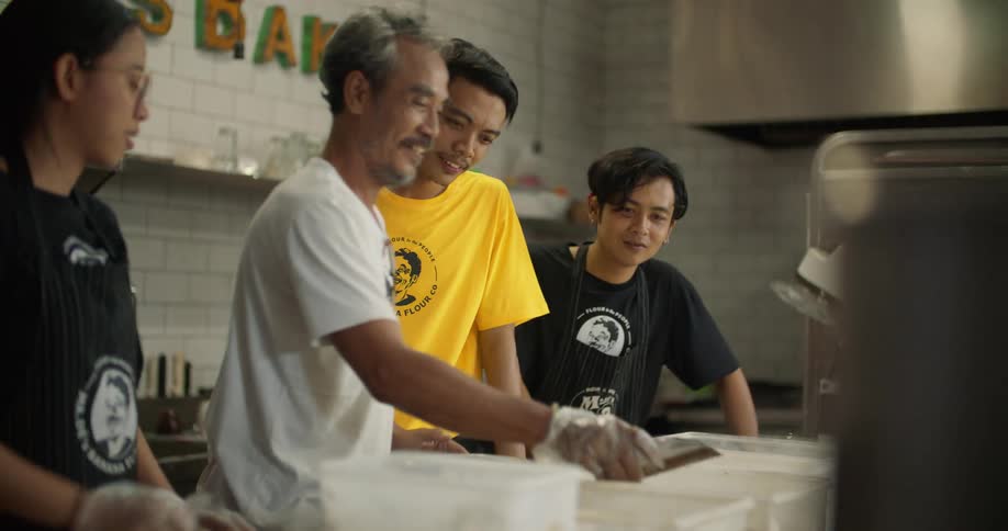 A man uses a scoop to add ingredients to bins in a bakery as his young employees look on