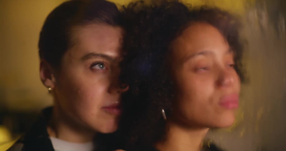A woman looking out of a window with water running down the surface is embraced from behind by another woman