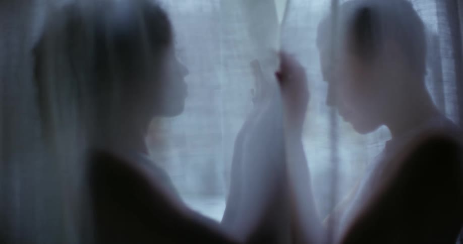 Two women are standing behind a sheer curtain facing each other as they hold their arms up and press their hands together