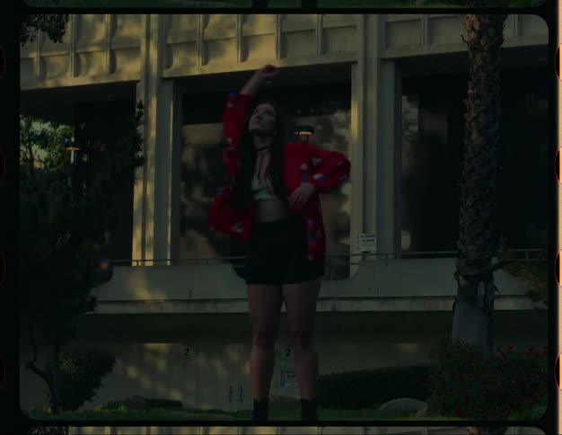 A teenage girl is standing outside of a building dancing near a tree