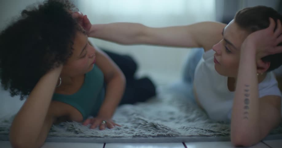 Two women lie on rug facing each other as one strokes the others head