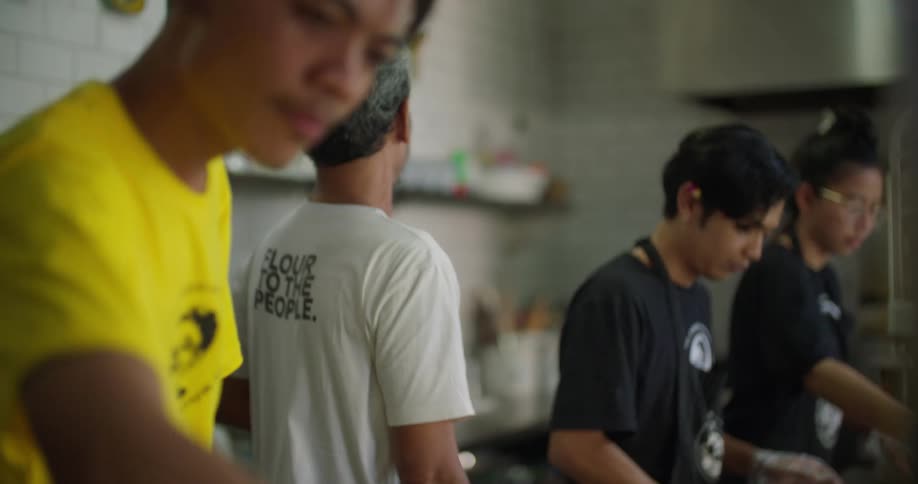 A group of young adults cook together in an industrial kitchen as an older man observes from behind