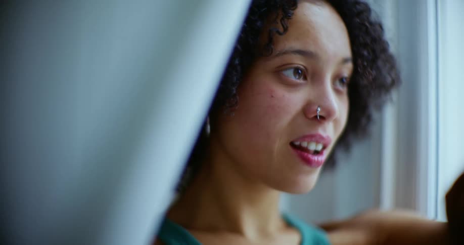 A woman blushes and smiles as she stands by a window holding back the sheers