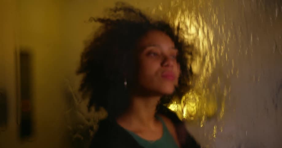 A young black woman is inside looking outside the window as it rains