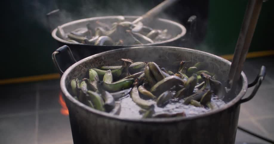 A pair of people stir boiling pots of bananas with sticks