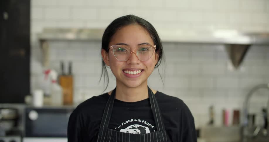 A young woman in an apron turns her head to smile into the camera while standing in a professional kitchen