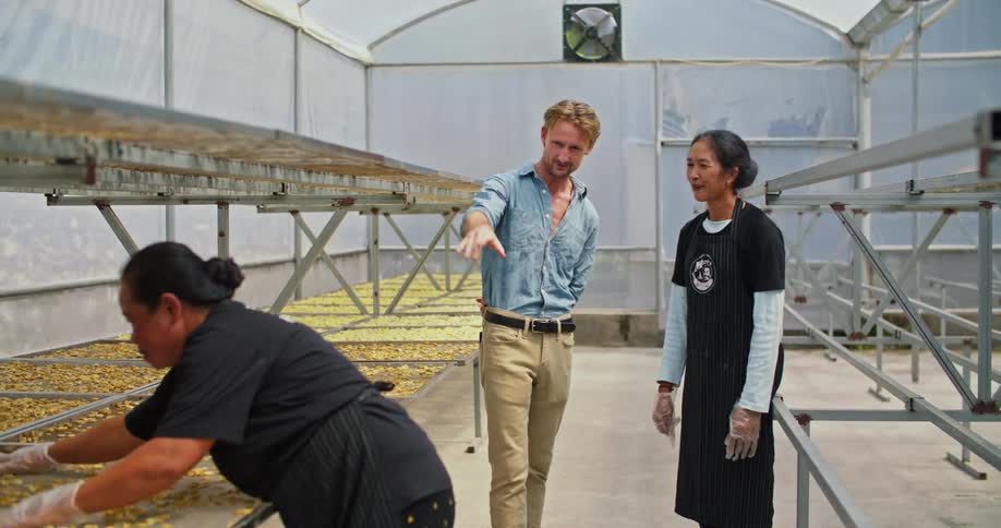 A man stands talking with a woman next to trays full of dehydrating banana chips