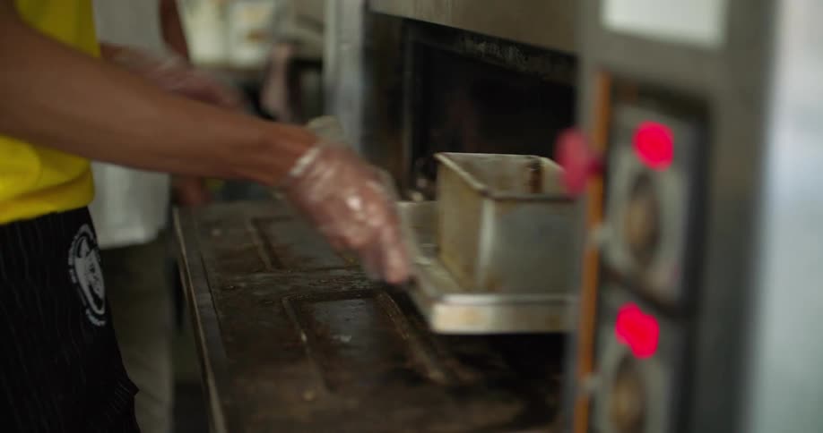A person puts a baking pan into a large industrial oven as another person observes from nearby