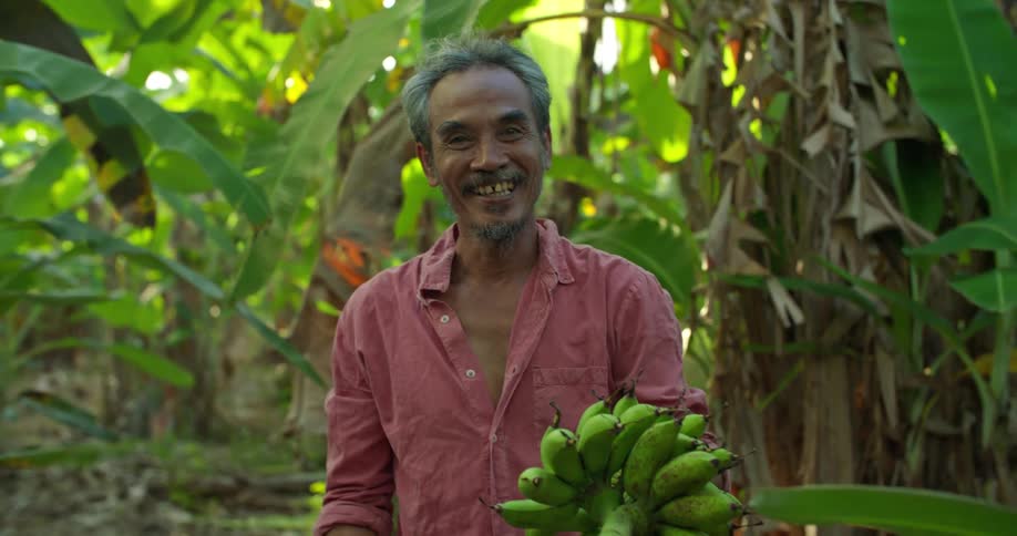 A man standing in a forest smiles into the camera while presenting a bunch of bananas