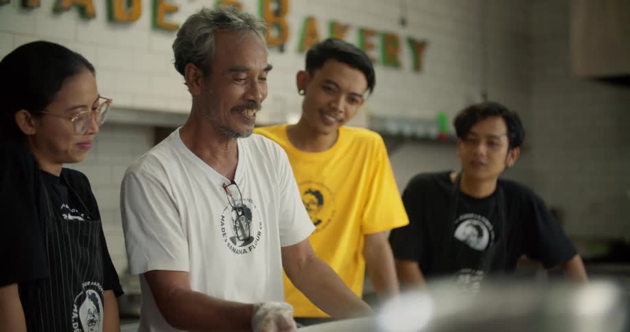 A man holding a plastic bin in a bakery talks and teaches his young workers