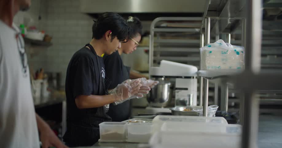 A man and woman prepare food in the kitchen of a restuarant as another man carries dishes behind them