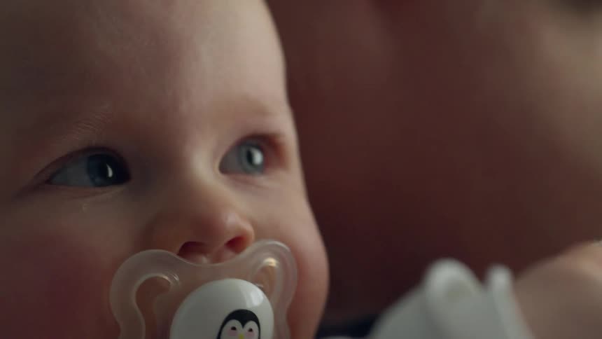 Baby with a pacifier in its mouth moves its hand from his head to the shoulder of an adult
