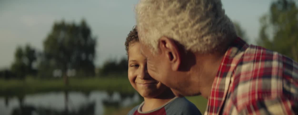 A boy is standing outside with his grandfather and they are looking at a pond as they talk