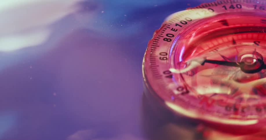 A compass with spinning needle is covered with water reflecting colorful light