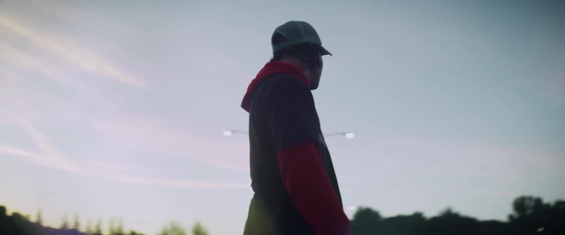 A teenage boy wearing a coat and cap is gliding across a parking lot on a skateboard and there are trees and streetlights nearby