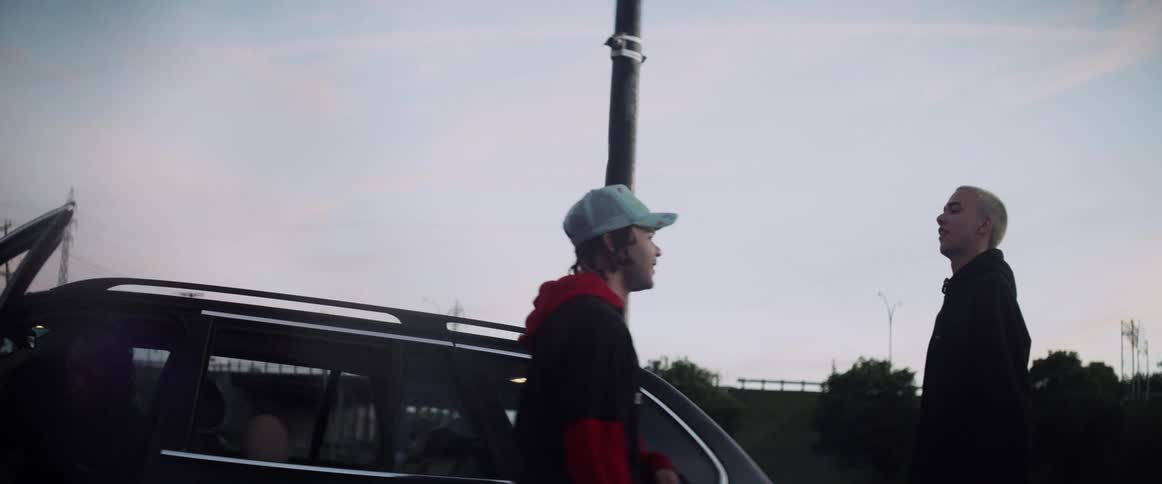 A teenage boy walks up to his friend standing by a car and laughs as he pushes him and they pretend to fight and laugh