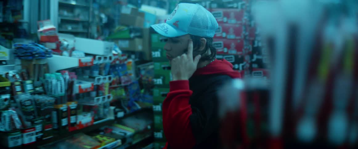 A teenage boy carries a drink through a store and sets it on a counter and leans over the counter to look for the cashier