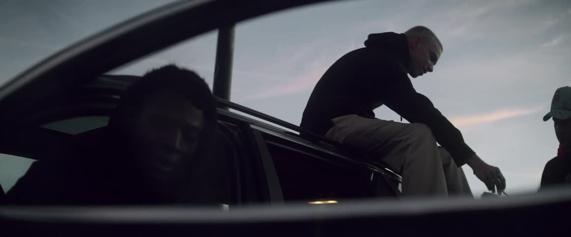 A man sits smoking in his car door open when friends approach one jumping onto the roof