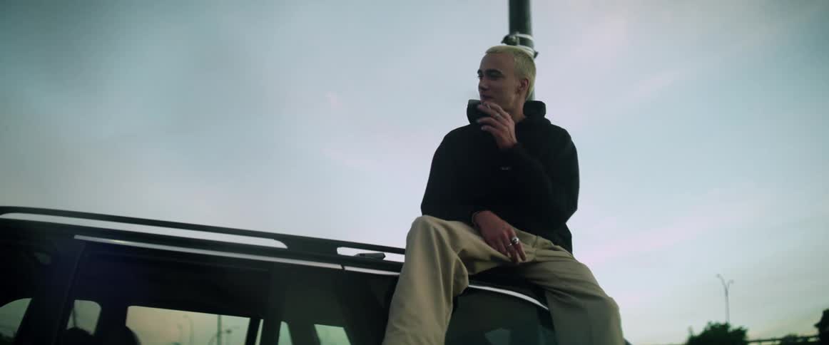 A man is sitting on the roof of an automobile smoking a cigarette as a teenage boy passes by