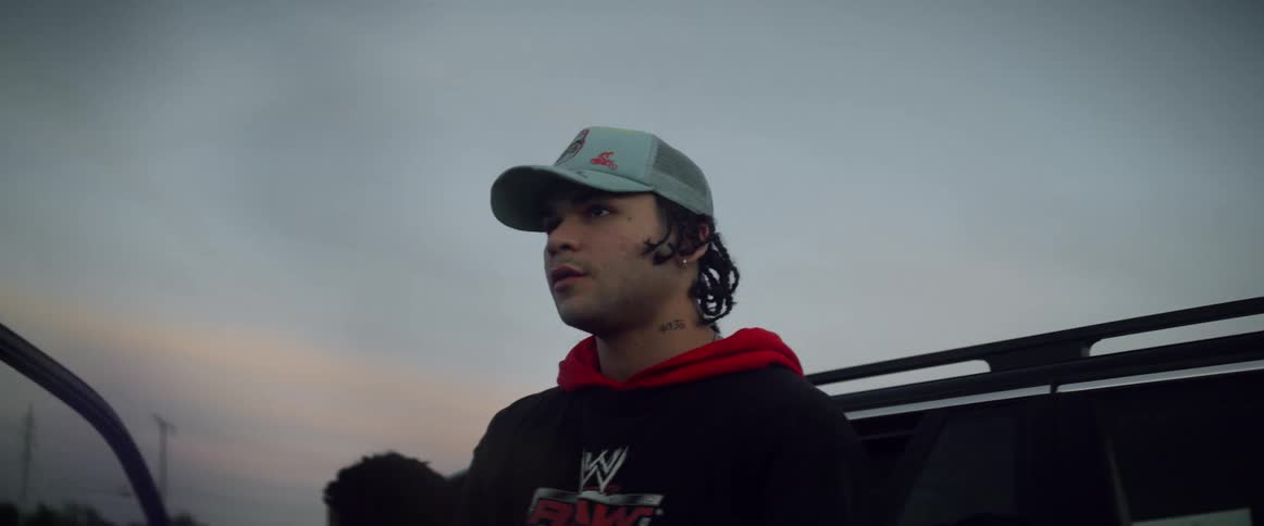 A teenage boy is sitting in the drivers seat of a parked vehicle smoking a cigarette and another boy is leaning against the side of the automobile