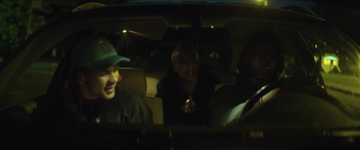Three teenagers are traveling in an automobile and the driver is keeping his eyes on the road while the two passengers are eating fries and playing around trying to distract him