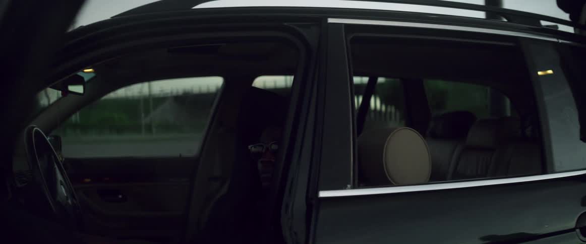 A man is sitting in the drivers seat of a parked automobile with the door open and a teenage boy walks around the vehicle and talks to a person sitting on the roof