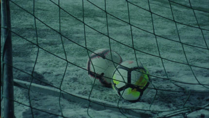 Two soccer balls sitting in the net on a snow dusted field
