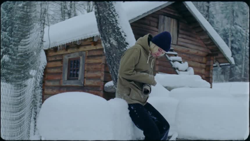 A sad looking teenage girl walks through deep snow by a log cabin and sits on a bench covered in snow and pouts