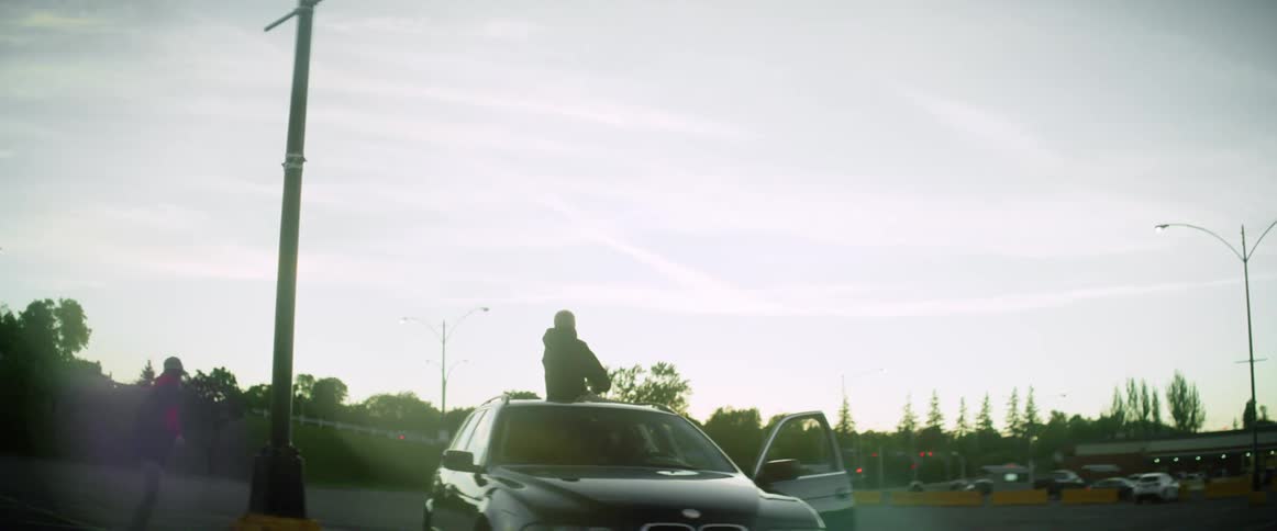 A teenage boy skateboards around a car his friend is sitting on and then jumps off his skateboard and puts his hands on the front of the car and flips over the hood
