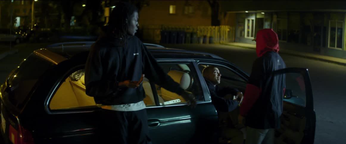 Two teenage boys stand outside a car and talk and point and then one of the boys tries to pull another boy that is sitting in the car out