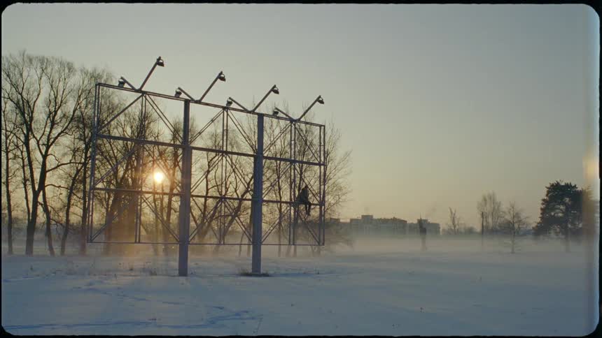 A teenager sitting on a sports field scoreboard bracket above the snow swinging her legs