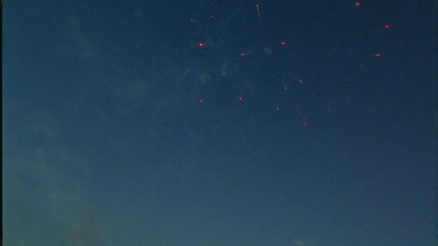 Three teenage boys are on the roof of a building shooting off fireworks and silhouettes of other buildings are in the distance