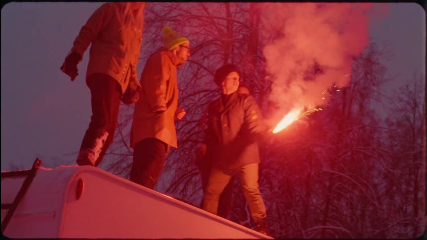 A group of friends dancing on the roof of an rv with a smoking red flare they throw into the snow
