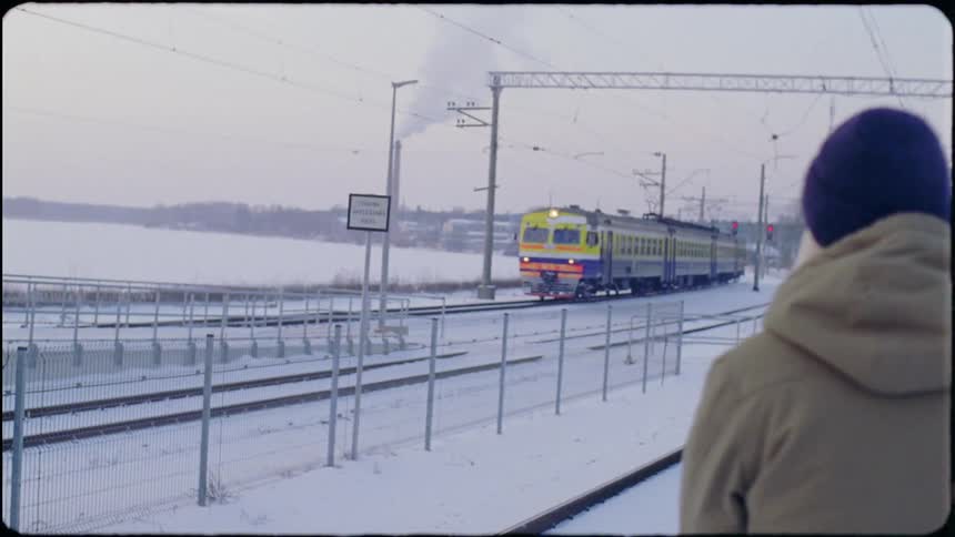 A woman with long blond hair wearing a knit cap and a coat is standing outside in the snow near train tracks as she watches a train pass
