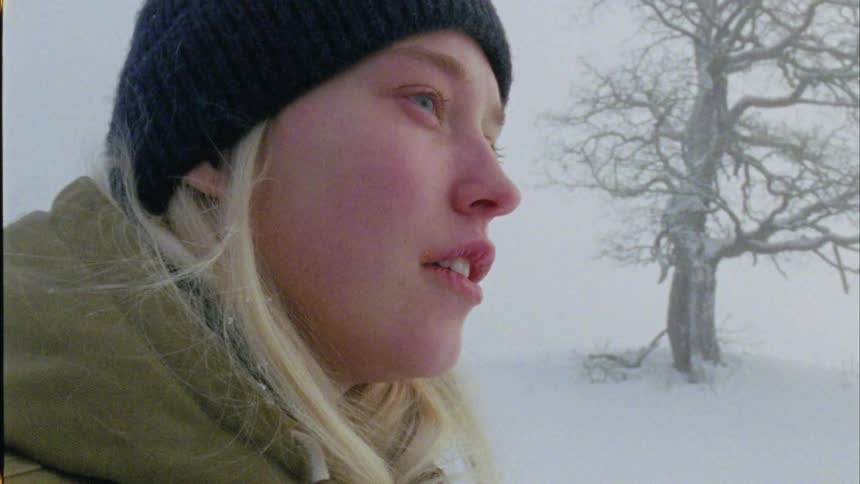 A teenage girl with long blond hair wearing a knit cap and a coat is standing outside in the snow near a tree as she wipes her face with her gloved hand