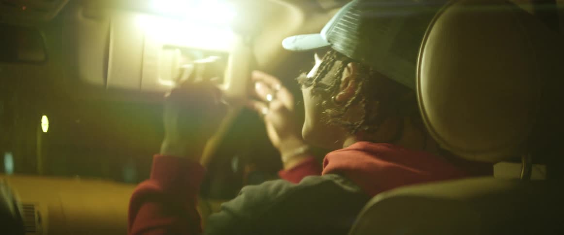A teenager in the passenger seat checks his hat in the mirror while smiling and talking